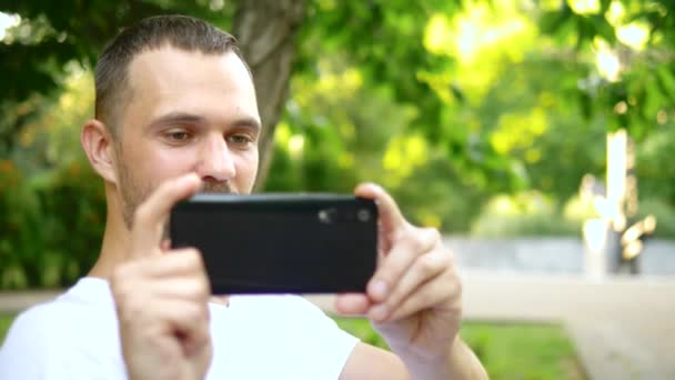 Primer plano. Un bloguero hombre utiliza un teléfono inteligente mientras está sentado en un banco del parque. Tiene una videollamada con alguien — Vídeos de Stock