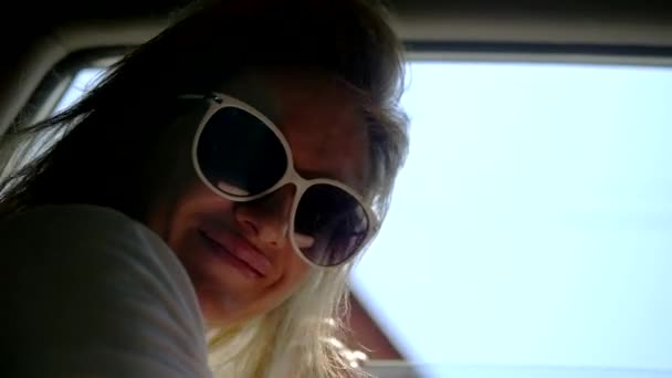 Счастливая молодая женщина в солнечных очках смотрит в открытое окно машины, и ее длинные волосы взволнованы ветром. Авто путешествия и приключения — стоковое видео