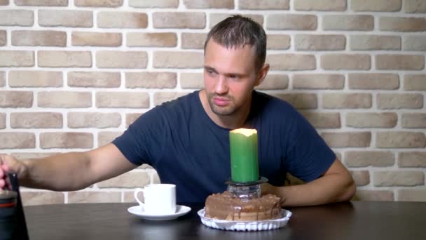 Narozeninový koncept pro jednotlivce, svobodný, osamělý člověk. mladý muž sedící sám u stolu proti cihlové zdi. před ním je čokoládový dort s obrovskou sváteční svíčkou — Stock video