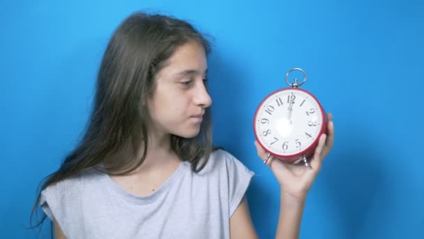 El concepto de tiempo de aprendizaje, de vuelta a la escuela. una colegiala sostiene un gran reloj y se sorprende de que haya llegado el momento de aprender. sobre fondo azul — Vídeos de Stock