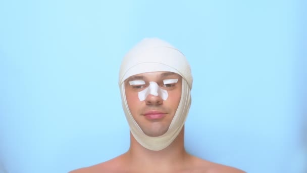 En man efter en plastikkirurgi Facelift, näsplastik, Ögonlocksplastik. med ett bandage på näsan, huvudet och ögonen. på blå bakgrund. visar en super Sign och okej — Stockvideo