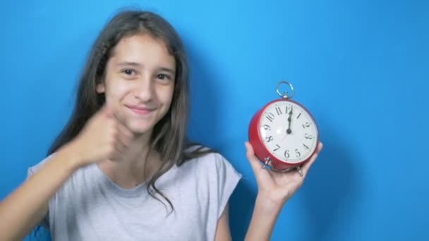 Het concept van leertijd, terug naar school. Een schoolmeisje houdt een grote klok die naar hen wijst en glimlacht dat het tijd is om te leren. op blauwe achtergrond — Stockvideo