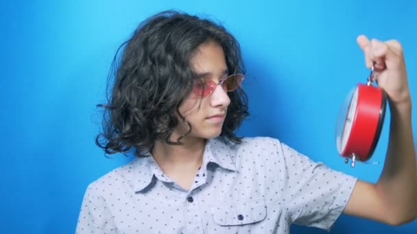 Adolescente drôle dans des lunettes roses tient une horloge dans ses mains, regarde l'heure et ferme son visage avec sa main . — Video