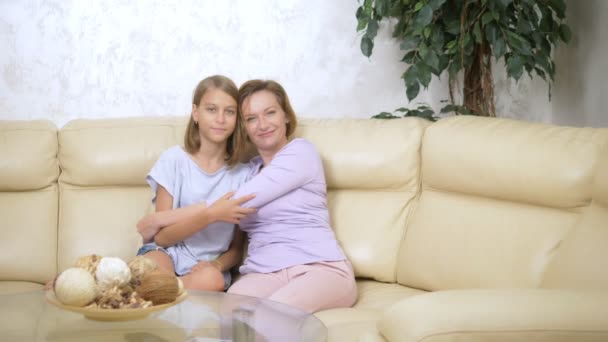Teenage girl and her mother hugging, looking at the camera and smiling while sitting on the sofa in the living room — Stock Video