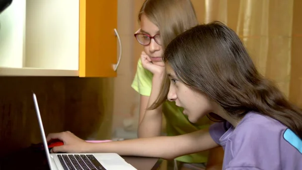 Δύο κοπέλες φιλενάδες, έφηβοι μαθητές κάθονται μαζί στο τραπέζι εκπαίδευσης και χρησιμοποιούν ένα φορητό υπολογιστή το βράδυ. είναι χαρούμενες και ευτυχισμένες — Φωτογραφία Αρχείου