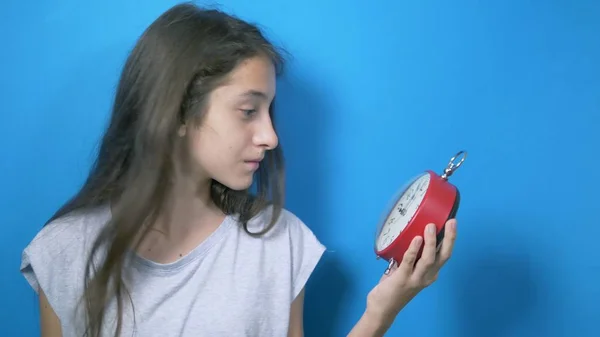 Das Konzept der Lernzeit, zurück zur Schule. Eine Schülerin hält eine große Uhr in der Hand und ist überrascht, dass die Zeit gekommen ist, zu lernen. auf blauem Hintergrund — Stockfoto