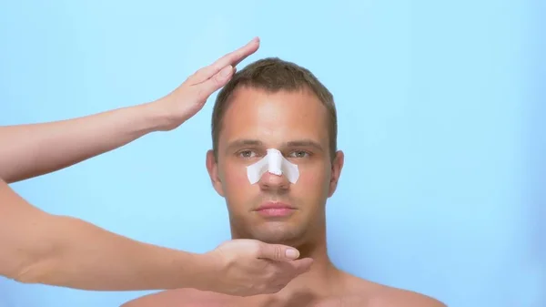 Концепция пластической хирургии, руки держат лицо человека. мужчина после пластической операции на лице, ринопластика, с повязкой на носу. на синем фоне . — стоковое фото