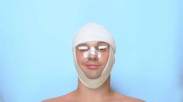 A man after a plastic surgery facelift, rhinoplasty, blepharoplasty. with a bandage on the nose, head and eyes. on blue background. shows a super sign and okay — Stock Photo, Image