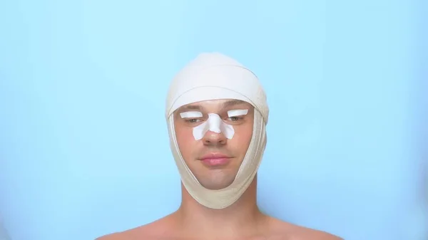 a man after a plastic surgery facelift, rhinoplasty, blepharoplasty. with a bandage on the nose, head and eyes. on blue background. shows a super sign and okay