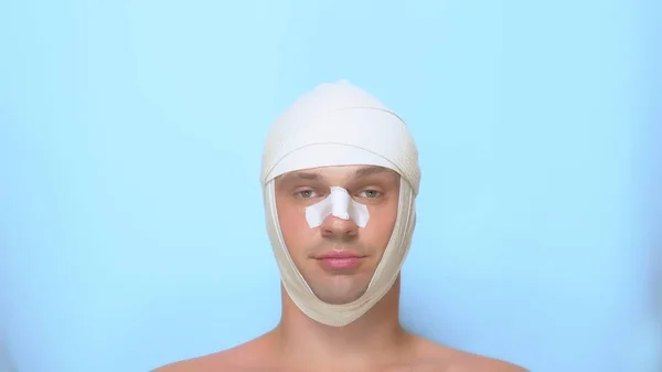 A man after a plastic surgery facelift, rhinoplasty, blepharoplasty. with a bandage on the nose, head and eyes. on blue background. shows a super sign and okay — Stock Photo, Image