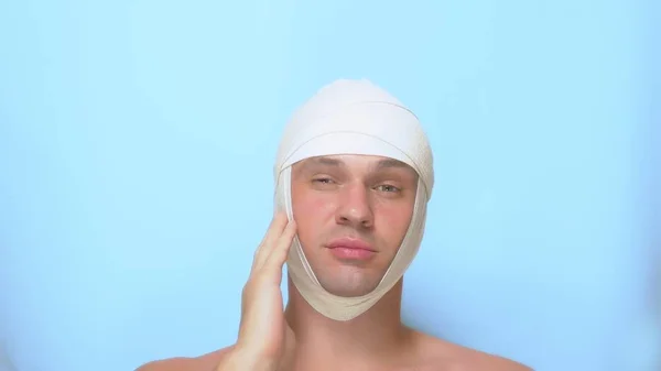 Het concept van plastische chirurgie. een man raakt zijn gezicht na een plastische chirurgie gezicht lift, met een verband op zijn hoofd. op blauwe achtergrond. — Stockfoto