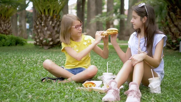 Μαθητές πικ-νικ. δύο κορίτσια γκόμενες τρώνε χάμπουργκερ και τηγανιτές πατάτες που κάθονται στο γρασίδι στο πάρκο — Φωτογραφία Αρχείου