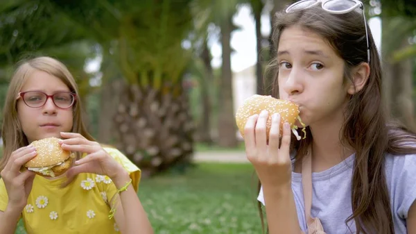 Konsep piknik siswa. dua gadis pacar makan hamburger dan kentang goreng duduk di rumput di taman — Stok Foto