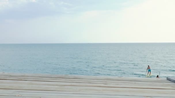 Pohled na dřevěnou terasu od desek k moři a k nebi. dva veslaři plavou na prkně z rámu. místo pro kopírování. mořské pozadí — Stock video
