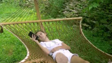 genç kız ormanda bir piknik bir ağacın altında bir hamakta yatarken bir akıllı telefon kullanır