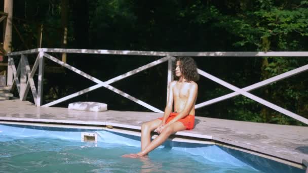 Sonriente adolescente con el pelo moreno rizado sentado junto a la piscina, las piernas abajo en el agua. vacaciones de verano — Vídeos de Stock