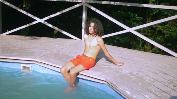 Усміхнений хлопчик-підліток з кучерявим волоссям брюнетки, що сидить біля басейну, ноги вниз у воді. літні канікули — стокове відео