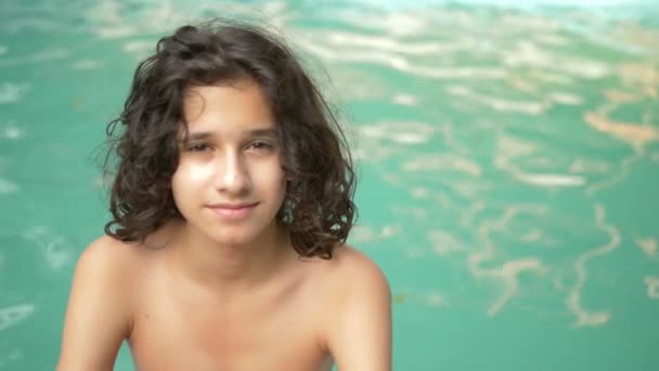 Garoto adolescente sorridente com cabelo morena encaracolado sentado à beira da piscina, brincadeira em um fundo de água azul — Vídeo de Stock