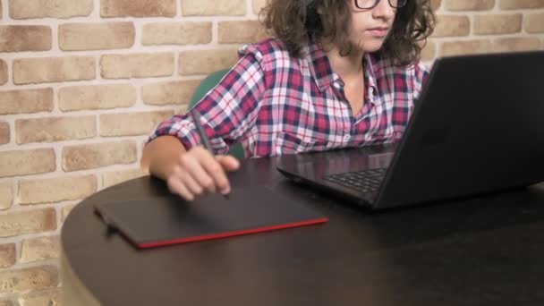 Cerca. adolescente con el pelo rizado morena, en una camisa a cuadros funciona en una tableta gráfica utilizando un lápiz — Vídeos de Stock