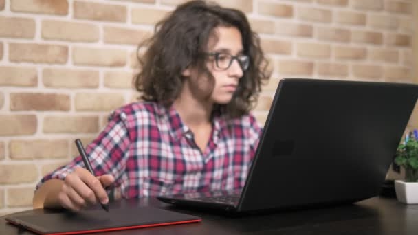 Nära. tonåring pojke med lockigt brunett hår, i en pläd skjorta fungerar på en grafik tablett med en penna — Stockvideo