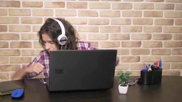 Guay adolescente con el pelo moreno rizado, en una camisa a cuadros, en auriculares, bailando, sentado frente a su computadora portátil, en una habitación tipo loft, contra una pared de ladrillo . — Vídeo de stock