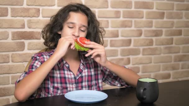 Adolescente hambriento con apetito come un sándwich con lechuga fresca y tomates en la cocina estilo loft contra una pared de ladrillo — Vídeos de Stock