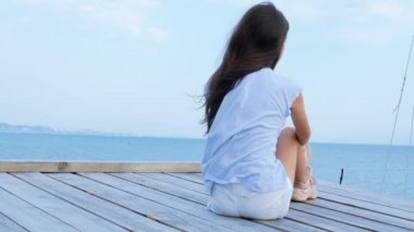Genç kız denizde ahşap bir köprü üzerinde tek başına oturur ve denizde üzgün görünüyor