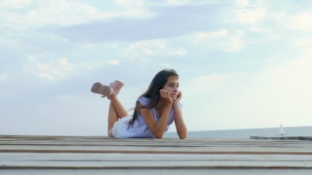 Tini lány ül egyedül egy fahíd a tengeren, és úgy néz ki, szomorú a tengeren — Stock videók