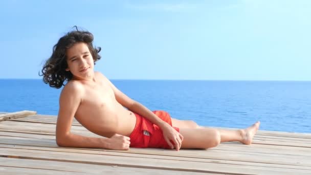 En vacker tonåring pojke med lockigt svart hår ligger på en träterrass ovanför havet. begreppet sommarlov, skollov — Stockvideo