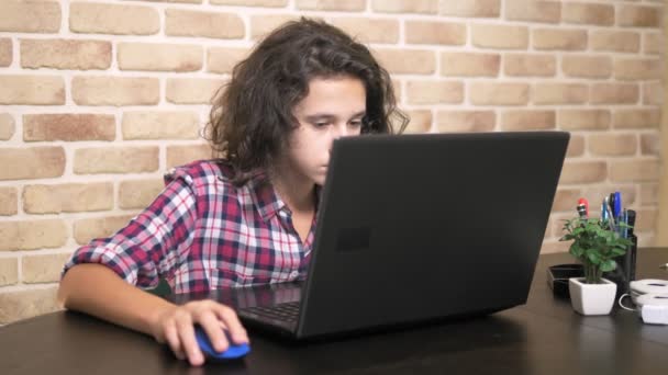 Dolgozik egy laptop számítógép, gépelés-ra egy billentyűzet, modern tinédzser. Egy tizenéves fiú göndör haja a kockás inget használ egy laptop egy loft stílusú szoba. — Stock videók