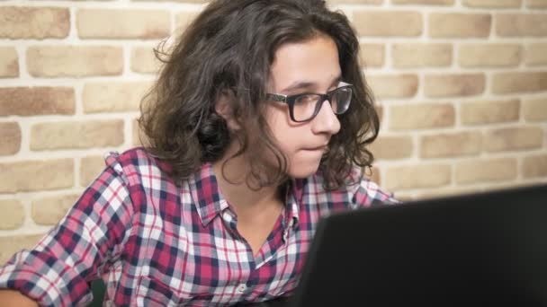 Trabajando en una computadora portátil, escribiendo en un teclado, adolescente moderno. Un adolescente con el pelo rizado en una camisa a cuadros utiliza un ordenador portátil en una habitación de estilo loft . — Vídeos de Stock