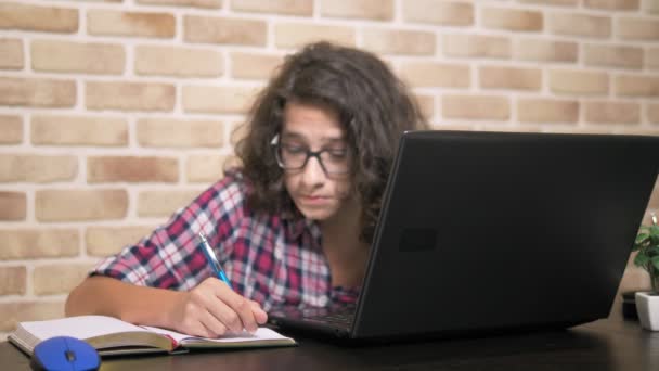 Hermoso adolescente con el pelo moreno rizado trabajando en su computadora portátil y escribiendo en su cuaderno, sentado en una mesa contra una pared de ladrillo. estilo de diseño loft — Vídeos de Stock
