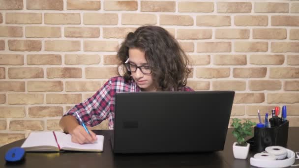 Красивый мальчик-подросток с вьющимися брюнетками работает над ноутбуком и пишет в блокноте, сидя за столом у кирпичной стены. Лофт дизайн стиль — стоковое видео