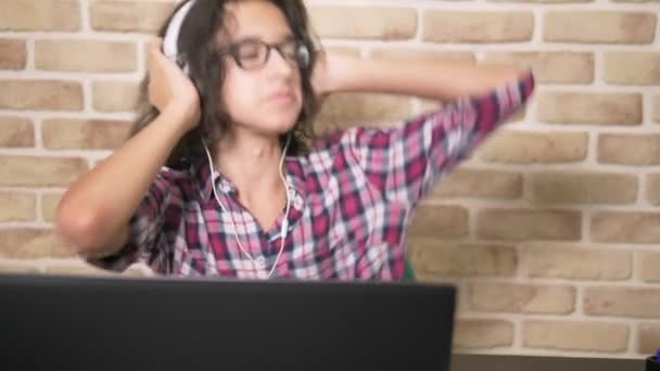 Крутой подросток с вьющимися брюнетками, в клетчатой рубашке, в наушниках, танцует, сидит перед ноутбуком, в лофт-комнате, напротив кирпичной стены . — стоковое видео