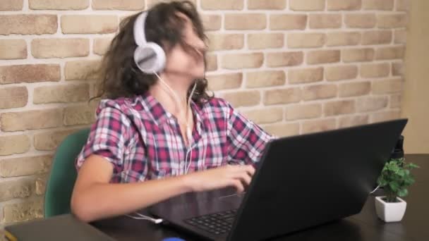 Coole tiener jongen met krullend brunette haar, in een geruit overhemd, in een koptelefoon, dansen, zittend in de voorkant van zijn laptop, in een kamer in loft-stijl, tegen een bakstenen muur. — Stockvideo