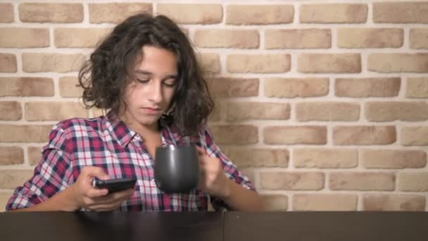 Hungry teen boy eats in the kitchen and uses a smartphone. — Stock Video