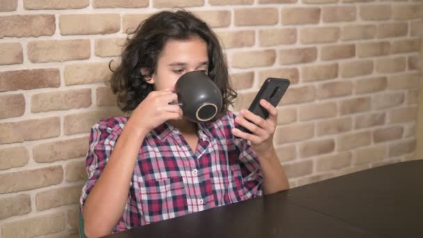 Fome adolescente menino come na cozinha e usa um smartphone . — Vídeo de Stock