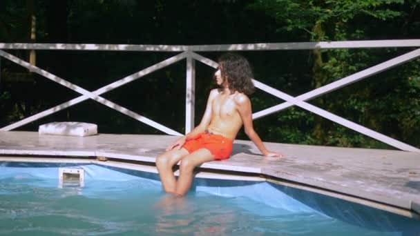 Adolescent souriant avec des cheveux bruns bouclés assis près de la piscine, les jambes dans l'eau. vacances d'été — Video