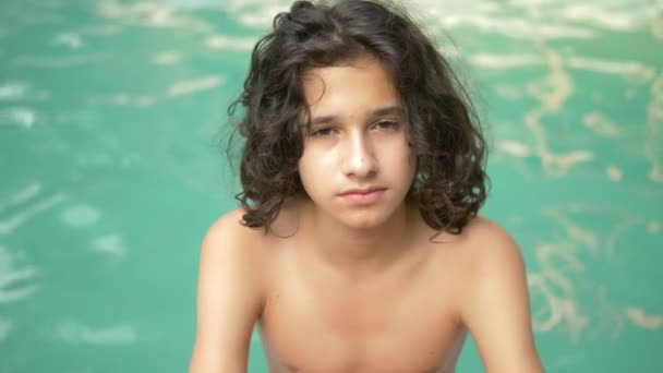 Ragazzo adolescente sorridente con i capelli ricci bruna seduto a bordo piscina, prtret su uno sfondo di acqua blu — Video Stock