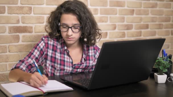 Bellissimo ragazzo adolescente con i capelli ricci bruna che lavora sul suo computer portatile e scrive nel suo quaderno, seduto a un tavolo contro un muro di mattoni. stile loft design — Video Stock