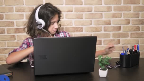 Guay adolescente con el pelo moreno rizado, en una camisa a cuadros, en auriculares, bailando, sentado frente a su computadora portátil, en una habitación tipo loft, contra una pared de ladrillo . — Vídeos de Stock