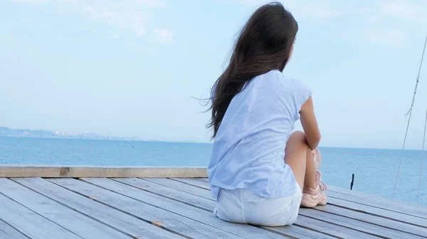 Genç kız denizde ahşap bir köprü üzerinde tek başına oturur ve denizde üzgün görünüyor — Stok fotoğraf