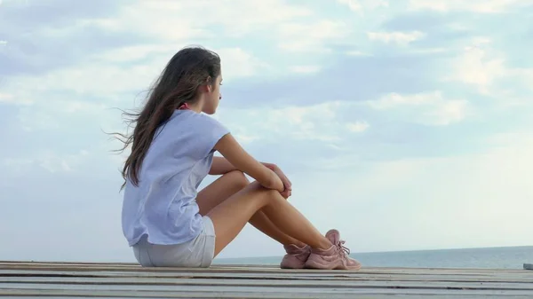 Adolescente chica se sienta sola en un puente de madera en el mar y se ve triste en el mar —  Fotos de Stock