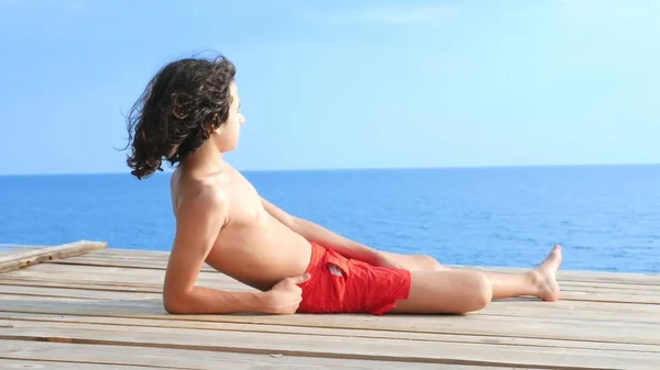 巻き毛の黒い髪をしたハンサムなティーンエイジャーの少年は、海の上の木製のテラスに横たわっています。夏休み、学校休業の概念 — ストック写真