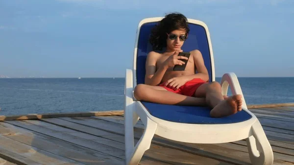 Kıvırcık siyah saçlı güzel genç çocuk deniz üzerinde ahşap bir terasta bir şezlong üzerinde yatıyor. akıllı telefon kullanır. yaz tatili kavramı, okul tatilleri — Stok fotoğraf