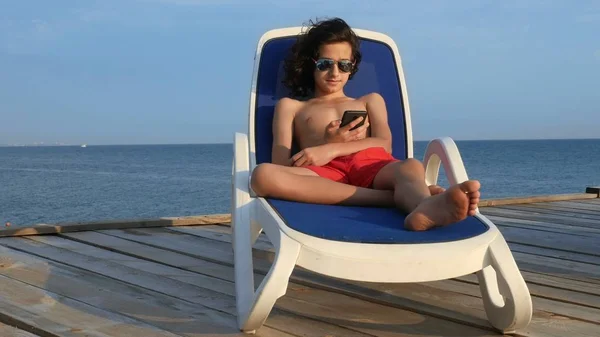O menino adolescente bonito com cabelo preto encaracolado encontra-se em uma cadeira de plataforma em um terraço de madeira sobre o mar. usa um smartphone. conceito de férias de verão, férias escolares — Fotografia de Stock