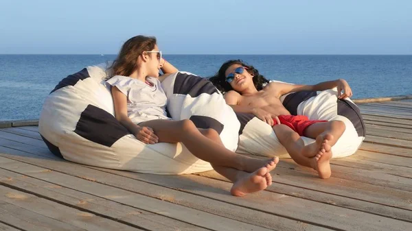 Belo adolescente menino e menina sentado em uma cadeira saco em um terraço de madeira sobre o mar. Eles falam alegremente. conceito de férias de verão, férias escolares — Fotografia de Stock