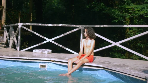 Havuz başında oturan kıvırcık esmer saçlı gülümseyen genç çocuk, bacaklar aşağı suda. yaz tatilleri — Stok fotoğraf