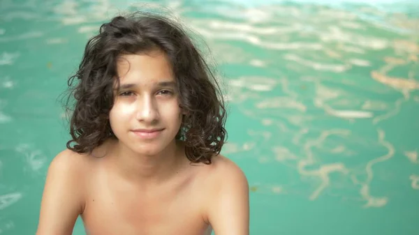 Lächelnder Teenager mit lockigen brünetten Haaren am Pool sitzend, prtret auf einem Hintergrund aus blauem Wasser — Stockfoto
