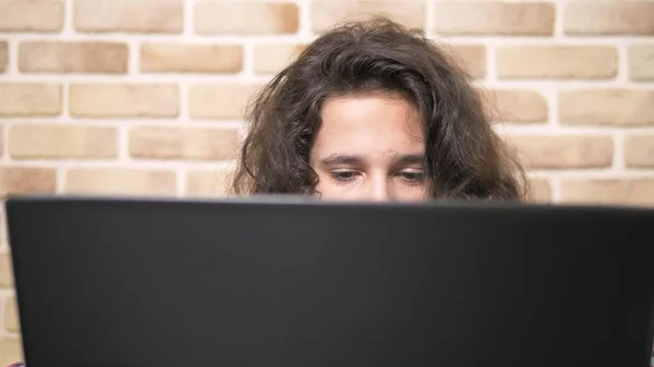 Praca na komputerze przenośnym, wpisując na klawiaturze, nowoczesny nastolatek. Nastoletni chłopiec z kręconymi włosami w koszulce w kratę używa laptopa w pokoju w stylu loftu. — Zdjęcie stockowe